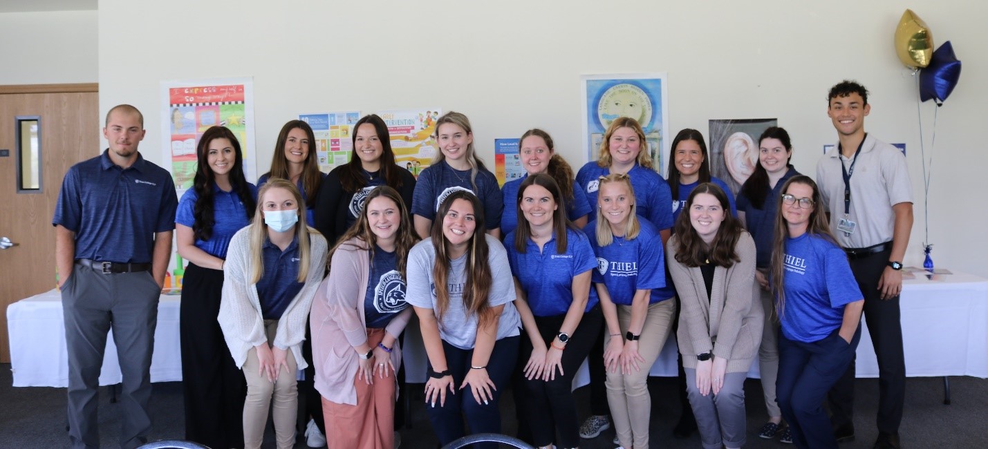Seventeen graduate students from the Thiel College Speech-Language Pathology program stand together in front of educational tables at the open house.
